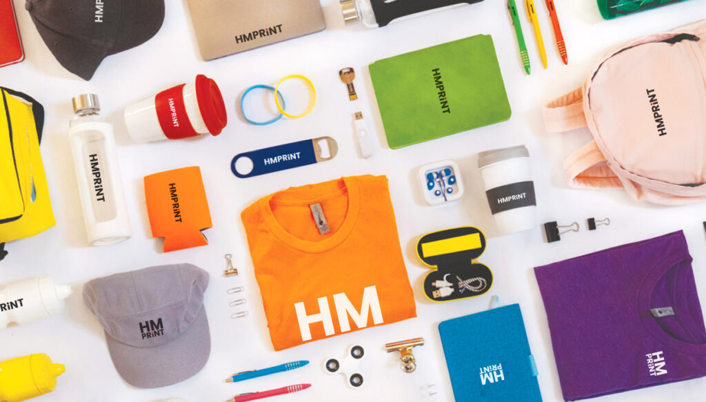 A variety of branded promotional gifts including hats, t-shirts, water bottles, and notebooks with the HMPRINT logo.